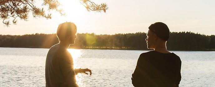 Två personer samtalar. De står vid en sjö.